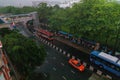 Vehicles on the street surrounded by trees and people standing under bus stop shelter during rain