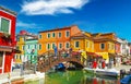 View on water canal with boats, wood bridge tre ponti, tourist crowd, gaudy brightly colored houses against blue sky, fluffy cloud