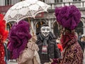Venice Carnival. A man in Guy Fawkes mask is posing with a costumed masked woman with a parasol and a costumed masked man