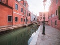 Venice, Italy - 17.10.2023: Calm water of a small canal with small bridge and red color buildings. Sun flare. Soft and airy mood.
