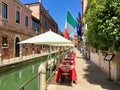 Tables and umbrellas of a restaurant set out along a quiet canal road with the Italian flag above