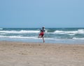 Male lifeguard wearing a sweatshirt and holding an orange life float running along a beach by the ocean