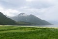 A verdant meadow leads to a fjord flanked by towering peaks shrouded in mist, encapsulating the serene beauty of the Norwegian