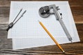 A vernier caliper holding a bearing, a pencil and a pair of compasses lying over drafting paper on wood background.