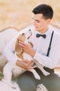 The vertical above portrait of the groom in vintage suit is petting the little dog and sitting on the white sofa in the