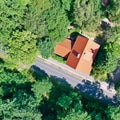Vertical aerial view of a house with a saddle roof and red roof tiles in a wooded area next to an asphalted grey country road