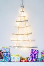Vertical background of Unusual Christmas tree made of wooden sticks with festive lights on the white wall and colourful gift boxes