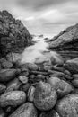 A vertical black and white sunset seascape photograph of misty waves crashing on the rocks