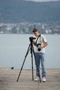 Vertical of a Caucasian guy on a wooden pier while shooting with a professional camera on a tripod Royalty Free Stock Photo