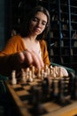 Vertical close-up portrait of young woman in elegant eyeglasses making chess move sitting on armchair in dark room Royalty Free Stock Photo