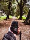 Vertical closeup of a human hand holding a black camera on a beautiful pathway background