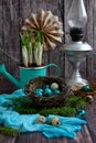 Vertical Easter composition. Painted quail eggs in a nest, plants in a decorative watering can and a kerosene lamp on a dark