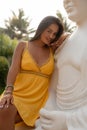 Vertical flirting, playful brunette woman in yellow dress sit on white plaster statue, look camera against green plants