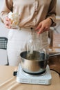 Vertical front view of female artisan adding white dry soy wax in glass jar into pot of boiling water for creating