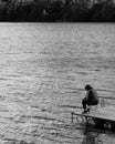 Vertical grayscale of a person fishing with a cart sitting on a deck on water