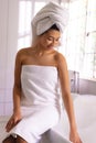 Vertical of happy biracial woman wearing towel sitting on the edge of bath smiling, with copy space Royalty Free Stock Photo