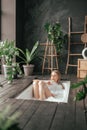 Vertical image of beautiful blonde young woman taking bath with bubbles in bathroom interior with plenty green plans