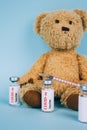 Vertical image of teddy bear with vial dose of COVID-19 vaccine with syringe against blue background with copy space -