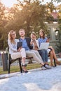 Vertical image of young caucasian adults laughing at bench in a park with their cell phones, laptop, tablet.  Looking at camera, Royalty Free Stock Photo
