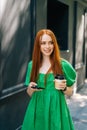 Vertical medium shot portrait of attractive young woman in green dress holding takeaway coffee cup, using mobile phone