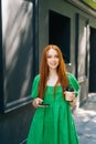 Vertical medium shot portrait of attractive young woman in green dress holding takeaway coffee cup, using mobile phone
