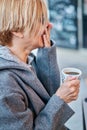 Vertical photo of Caucasian blonde woman wearing sunglasses sitting in a bar terrace with coat having a cup of coffee Royalty Free Stock Photo