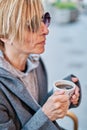 Vertical photo of Caucasian blonde woman wearing sunglasses sitting in a bar terrace with coat having a cup of coffee Royalty Free Stock Photo