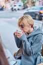 Vertical photo of Caucasian blonde woman wearing sunglasses sitting in a bar terrace with coat having a cup of coffee Royalty Free Stock Photo
