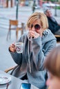 Vertical photo of Caucasian blonde woman wearing sunglasses sitting on a terrace of a bar with a coat having a cup of coffee with Royalty Free Stock Photo