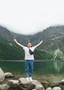 Vertical photo. Happy young man in casual clothes stands on a rock on the lake in the mountains with his hands up and enjoying the