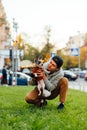 Vertical photo of a joyful funny young male dog-owner holding his cute doggy - york terrier, sitting on a green lawn near the busy Royalty Free Stock Photo