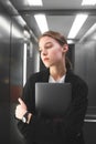 Vertical photo of serious young businesswoman holding her laptop in the elevator. Thoughtful calm female office worker is in the