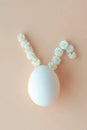 vertical photo of top view of white egg with ears like Easter bunny made from white flowers on peach fuzz background