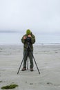 Vertical of a photographer standing in front of the camera on a tripod at the sandy beach Royalty Free Stock Photo