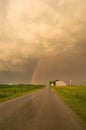 Vertical picture of a road surrounded by a field under a cloudy sky and a rainbow during the sunset Royalty Free Stock Photo