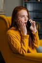 Vertical portrait of amazed excited young woman talking on smartphone sitting in yellow chair at home. Shocked female Royalty Free Stock Photo