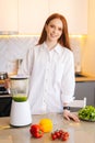 Vertical portrait of attractive redhead young woman making healthy vegetable detox smoothie juice in blender standing in Royalty Free Stock Photo