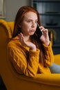 Vertical portrait of baffled young woman talking on smartphone sitting in yellow chair at home. Pretty shocked lady Royalty Free Stock Photo