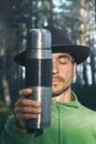 Vertical portrait of bearded millennial man in felt hat with closed eyes holding stainless thermos near his face on woods