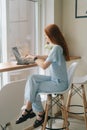 Vertical portrait of beautiful happy young woman typing on laptop computer sitting at table by window in cafe, looking Royalty Free Stock Photo