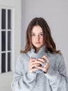 Vertical portrait of beautiful tween girl in warm grey pullover holding cup of tea looking at camera at home.