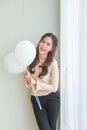 Vertical portrait beautiful young happy Asian woman in beige casual wear is smiling and holding white balloons side a white blinds Royalty Free Stock Photo