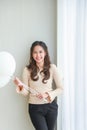 Vertical portrait beautiful young happy Asian woman in beige casual wear is smiling and holding white balloons side a white blinds Royalty Free Stock Photo
