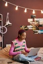 Black teenage girl sitting on bed with legs crossed using laptop at home Royalty Free Stock Photo