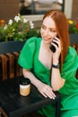 Vertical portrait of charming smiling young woman talking by mobile phone, sitting at table with coffee cup in outdoor Royalty Free Stock Photo