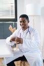 Vertical portrait of cheerful African-American male doctor wearing white uniform holding syringe in hand sitting at Royalty Free Stock Photo