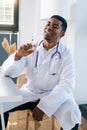 Vertical portrait of cheerful African-American male doctor wearing white uniform holding syringe in hand sitting at Royalty Free Stock Photo