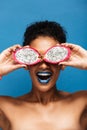 Vertical portrait of cheerful african american woman fooling around while covering eyes with exotic pitahaya fruit cut in half is