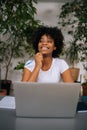 Vertical portrait of cheerful curly African female freelancer holding pen in hand, laughing looking at camera sitting at Royalty Free Stock Photo