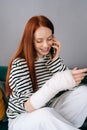 Vertical portrait of cheerful young woman injured hand wrapped in gypsum bandage talking on smartphone with friends